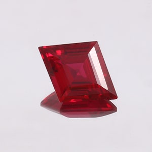 May include: A deep red, square-shaped gemstone with a faceted cut. The gemstone is set against a white background.