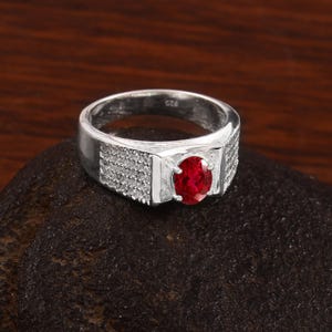 May include: A silver ring with a large, oval-cut, bright red gemstone at the centre. The band is wide and features small, clear stones on the sides. The ring is set against a dark brown background.