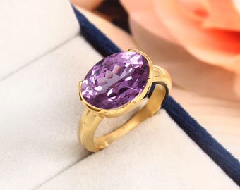 以下が含まれることがあります： 大きな楕円形の紫水晶をセットした金の指輪。指輪はシンプルなバンドに、石の周りに装飾的なデザインが施されています。