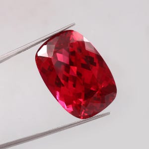 May include: A faceted, cushion-cut ruby gemstone held by tweezers. The ruby is a deep, vibrant red color with multiple facets that reflect light. The gemstone is rectangular with rounded corners.