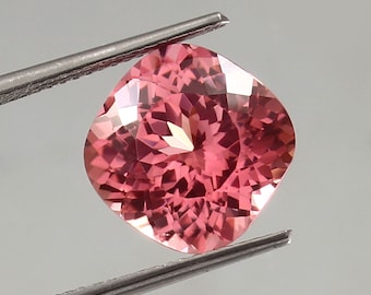 May include: A pink cushion-cut gemstone with a faceted surface. The gemstone is held by a pair of tweezers.