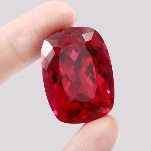 May include: A large, deep red gemstone with a cushion cut. The gemstone is held in a hand, showcasing its size and brilliance.
