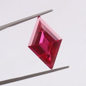 May include: A vibrant red gemstone with a square shape and a diamond-like cut. The gemstone is held by tweezers against a white background.