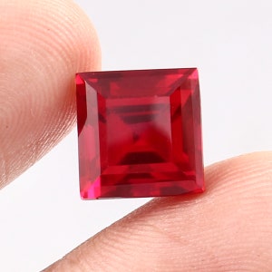 May include: A square-cut, vibrant red gemstone. The gem is held between fingers against a white background. The facets of the stone reflect light, showcasing its clarity and rich colour. The gemstone is a deep, saturated red.