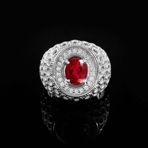 May include: A silver ring with a large oval, deep red gemstone at the center, surrounded by a halo of small, clear gemstones. The ring's band and setting are intricately detailed with a textured design.
