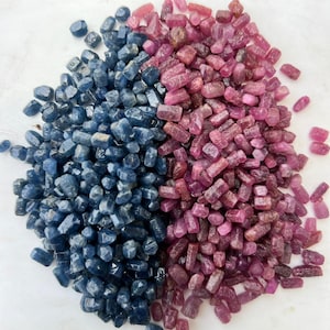 Peut inclure: Une grande collection de petites pierres précieuses brutes et polies, divisées en deux tas distincts. Un tas se compose de cristaux de saphir bleu profond, tandis que l'autre présente des cristaux de rubis rose et rouge violacé.