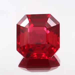 May include: A faceted, rectangular-cut ruby gemstone. The gem is a deep, saturated red color with a high degree of clarity. The facets create a brilliant display of light and reflection. The gemstone is set against a white background.