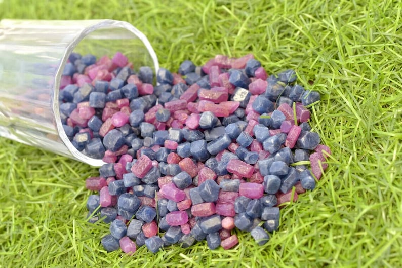 K&ouml;nnte beinhalten: Ein umgest&uuml;rzter, klarer Glasbeh&auml;lter, der eine Sammlung kleiner, polierter Edelsteine auf eine gr&uuml;ne Grasfl&auml;che verstreut. Die Edelsteine sind eine Mischung aus tiefblauen und leuchtend rosa Farbt&ouml;nen, was einen auff&auml;lligen Kontrast erzeugt.