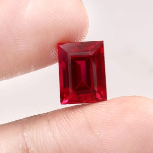 Könnte beinhalten: Ein rechteckig geschliffener Rubin in tiefroter Farbe. Der Edelstein wird zwischen Fingern gehalten, wodurch seine facettierte Oberfläche und innere Struktur sichtbar werden. Die rechteckige Form betont die Klarheit und Farbe.