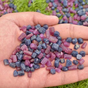 K&ouml;nnte beinhalten: Nahaufnahme einer Hand, die eine Sammlung roher Edelsteine in tiefblauen und magentafarbenen T&ouml;nen h&auml;lt. Die ungeschliffenen Steine liegen auf einer gr&uuml;nen Oberfl&auml;che.