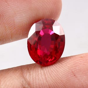 Könnte beinhalten: Ein facettierter, ovalförmiger Rubin-Edelstein. Der Edelstein hat eine tiefe, leuchtende rote Farbe mit mehreren Facetten, die das Licht reflektieren. Der Rubin wird zwischen zwei Fingern vor einem weißen Hintergrund gehalten.
