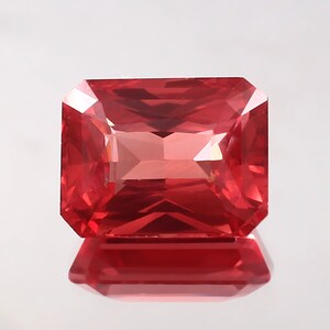 May include: A vibrant red gemstone with a rectangular cut and a polished finish. The gemstone is reflecting light, showcasing its brilliance.