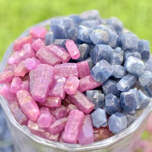 K&ouml;nnte beinhalten: Ein klares Glasgef&auml;&szlig; gef&uuml;llt mit rohen Edelsteinen. Die Edelsteine sind in zwei Bereiche unterteilt: rosa/rote Rubine und blaue Saphire. Die Edelsteine haben eine raue, nat&uuml;rliche Textur, vor einem verschwommenen gr&uuml;nen Hintergrund.
