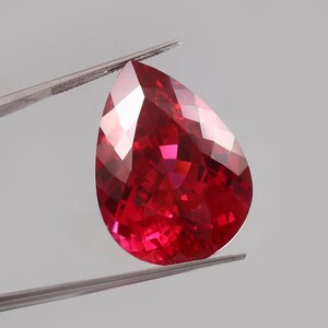 May include: A faceted, pear-shaped ruby gemstone. The gem is a deep red color with multiple facets that reflect light. Held by a pair of silver tweezers, the ruby is set against a neutral background.