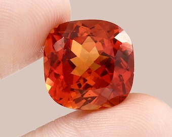 May include: A vibrant orange gemstone with a cushion cut. The gemstone has a star-like pattern in the center and is held in a hand.