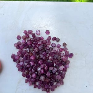 Może przedstawiać: Kolekcja małych, surowych kamieni szlachetnych rubinu w odcieniach od głębokiego różu do purpurowej czerwieni. Te luźne, nieoszlifowane kamienie mają nieregularne, fasetowane kształty o półprzezroczystym lub nieprzezroczystym wyglądzie, zebrane w gęstą stertę.