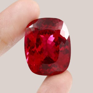 May include: A large, deep red gemstone with a cushion cut. The gemstone is held in a hand, and the background is a light grey.