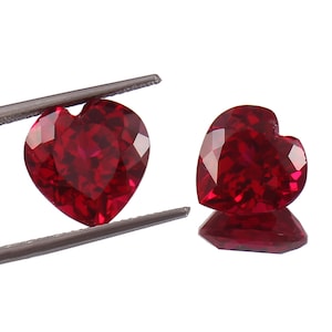 May include: Two heart-shaped red gemstones, likely rubies, are shown in a close-up image. The gemstones are held by tweezers and are displayed against a white background.