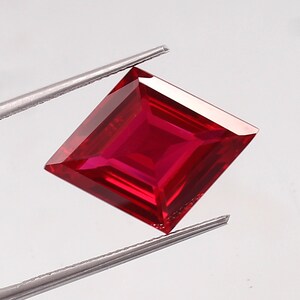 May include: A vibrant red square-shaped gemstone with a faceted cut. The gemstone is held by a pair of tweezers.