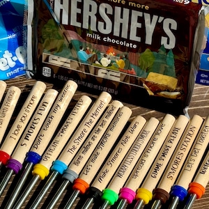 May include: A set of 12 wooden skewers with different s'mores-themed sayings. The skewers are made of wood and have a metal rod at the end. The sayings include "Life is smore fun with family", "I love you s'more", "S'mores Fun", "Don't touch my weenie", "Official S'mores Tester", "The S'more The Merrier", "Let's make S'more memories", "Make S'mores Not Wars", "Worry Less Laugh S'more", "Spread s'more love", "Gimme S'more Please", "S'mores Station", "Let's Get Toasted", and "Welcome to our firepit".