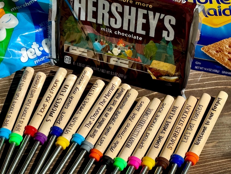 May include: A set of 12 wooden skewers with different s'mores-themed sayings. The skewers are made of wood and have a metal rod at the end. The sayings include "Life is smore fun with family", "I love you s'more", "S'mores Fun", "Don't touch my weenie", "Official S'mores Tester", "The S'more The Merrier", "Let's make S'more memories", "Make S'mores Not Wars", "Worry Less Laugh S'more", "Spread s'more love", "Gimme S'more Please", "S'mores Station", "Let's Get Toasted", and "Welcome to our firepit".