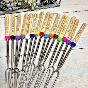May include: Set of 14 wooden handled s'mores roasting sticks with different engraved sayings. The sticks are silver and have a fork on the end. The sayings include "Spread s'more love", "Let's get toasted", "Worry less love s'more", "Don't touch my weenie", "Welcome to our firepit", "Make s'mores not wars", "Life is s'more fun with friends", "The s'more the merrier", "Gimme s'more please", "S'mores station", "Burnt mallows are the best mallows", "S'mores fun", "I love you s'more", "Official s'mores tester", and "Let's make s'more memories".