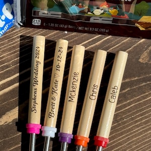 May include: Five wooden skewers with metal tips, each personalized with a name or date. The skewers are arranged in a row, with the tips facing the right. The skewers are on a wooden surface. The text on the skewers reads: "Stephens Wedding 2024", "Brett & Kate 10-12-24", "Mckenzie", "Chris", and "Caleb".