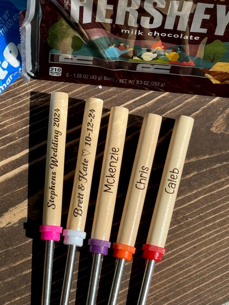 May include: Five wooden skewers with metal tips, each personalized with a name or date. The skewers are arranged in a row, with the tips facing the right. The skewers are on a wooden surface. The text on the skewers reads: "Stephens Wedding 2024", "Brett & Kate 10-12-24", "Mckenzie", "Chris", and "Caleb".