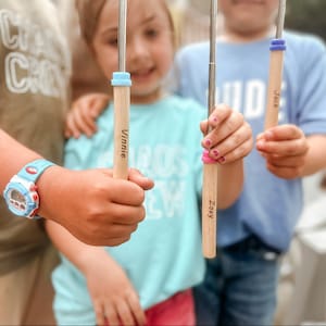 May include: Three children hold personalized wooden sticks with blue and purple accents. The sticks are labeled "Vinnie", "Zoey", and "Jack".