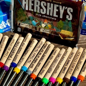 May include: A set of 12 wooden skewers with different s'mores-themed sayings. The skewers are arranged in a row on a wooden surface. The sayings include "Life is smore fun with family", "I love you s'more", "S'mores Fun", "Don't touch my weenie", "Official S'mores Tester", "The S'more The Merrier", "Let's make S'more memories", "Make S'mores Not Wars", "Worry Less Laugh S'more", "Spread s'more love", "Gimme S'more Please", "S'mores Station", "Let's Get Toasted", and "Welcome to our firepit".