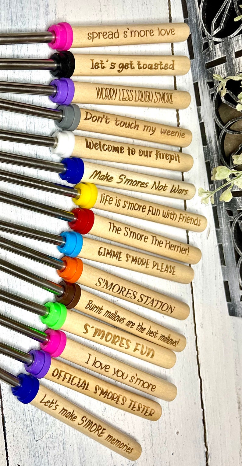 May include: A set of 12 wooden skewers with colorful tips and engraved sayings related to s'mores. The sayings include "spread s'more love", "let's get toasted", "worry less laugh smore", "don't touch my weenie", "welcome to our firepit", "make s'mores not wars", "life is s'more fun with friends", "the s'more the merrier", "gimme s'more please", "s'mores station", "burnt mallows are the best mallows", "s'mores fun", "i love you s'more", "official s'mores tester", and "let's make s'more memories".