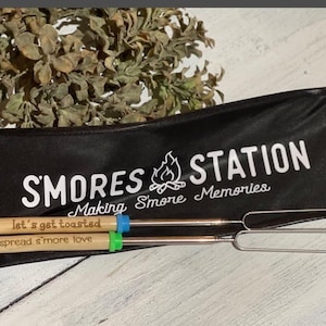 May include: A black storage bag with the white text SMORES STATION and Making S'more Memories, featuring two metal roasting sticks with wooden handles. One handle is engraved with let's get toasted and the other with spread s'more love.