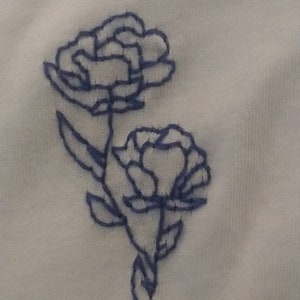 Puede incluir: Primer plano de tela blanca con bordado azul de flores. Se representan dos flores, con patrones detallados de p&eacute;talos y tallos. El bordado es en un azul contrastante, creando un delicado dise&ntilde;o floral.