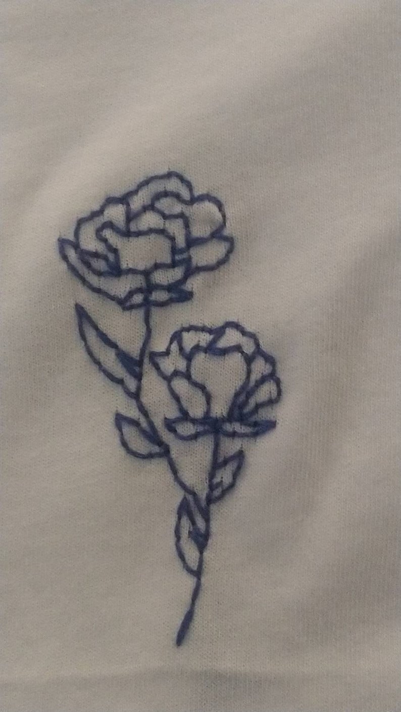 Puede incluir: Primer plano de tela blanca con bordado azul de flores. Se representan dos flores, con patrones detallados de p&eacute;talos y tallos. El bordado es en un azul contrastante, creando un delicado dise&ntilde;o floral.