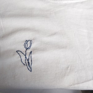 Puede incluir: Camiseta blanca lisa con cuello redondo y mangas cortas. Presenta un peque&ntilde;o dise&ntilde;o de tulip&aacute;n azul bordado en la parte delantera. El tulip&aacute;n tiene un estilo sencillo, con un tallo y hojas. La tela parece ser de algod&oacute;n suave.