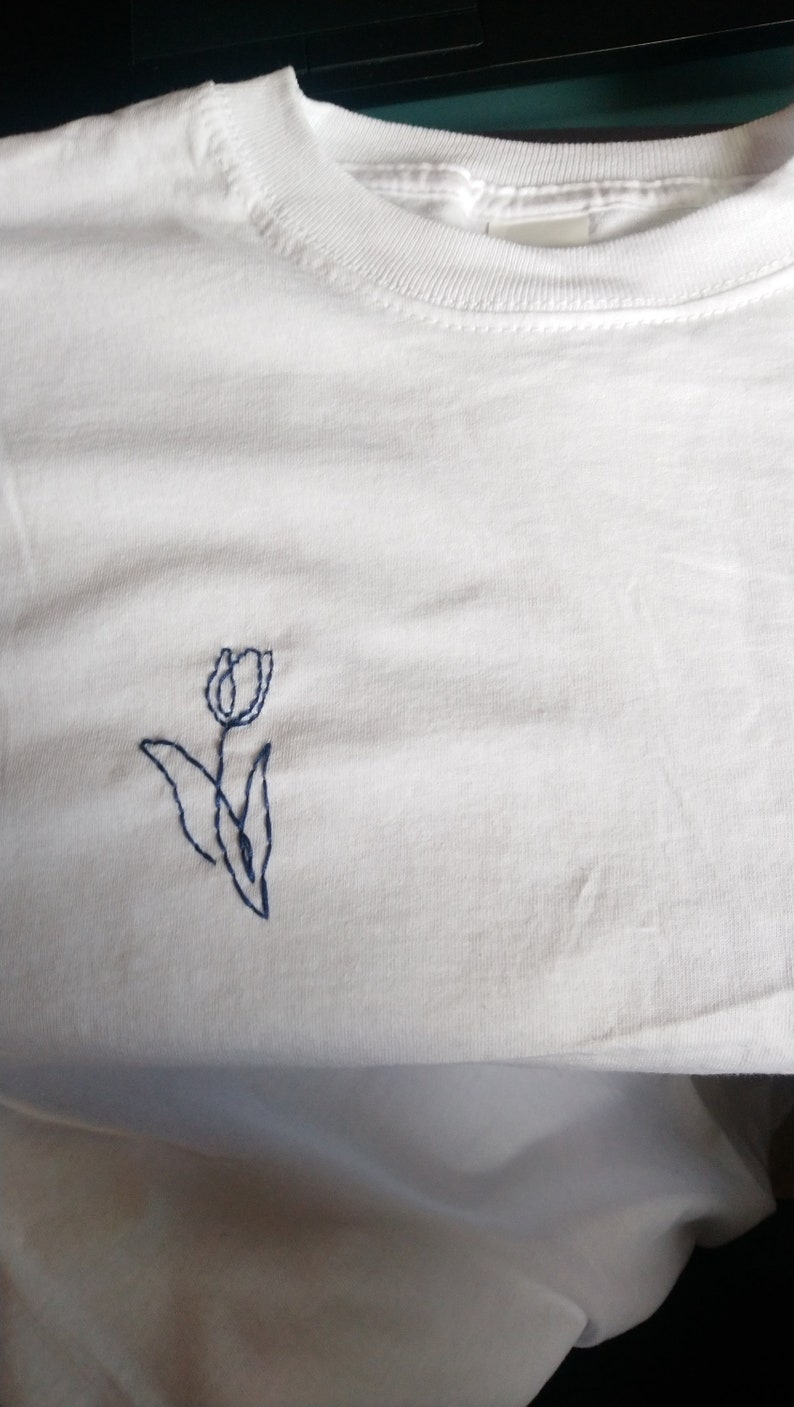 Puede incluir: Camiseta blanca lisa con cuello redondo y mangas cortas. Presenta un peque&ntilde;o dise&ntilde;o de tulip&aacute;n azul bordado en la parte delantera. El tulip&aacute;n tiene un estilo sencillo, con un tallo y hojas. La tela parece ser de algod&oacute;n suave.