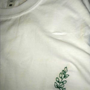 Puede incluir: Camiseta blanca con cuello redondo y mangas cortas. Un peque&ntilde;o dise&ntilde;o de hojas verdes bordadas est&aacute; en el pecho izquierdo. La camiseta est&aacute; hecha de un material suave. La etiqueta de la camiseta es visible.