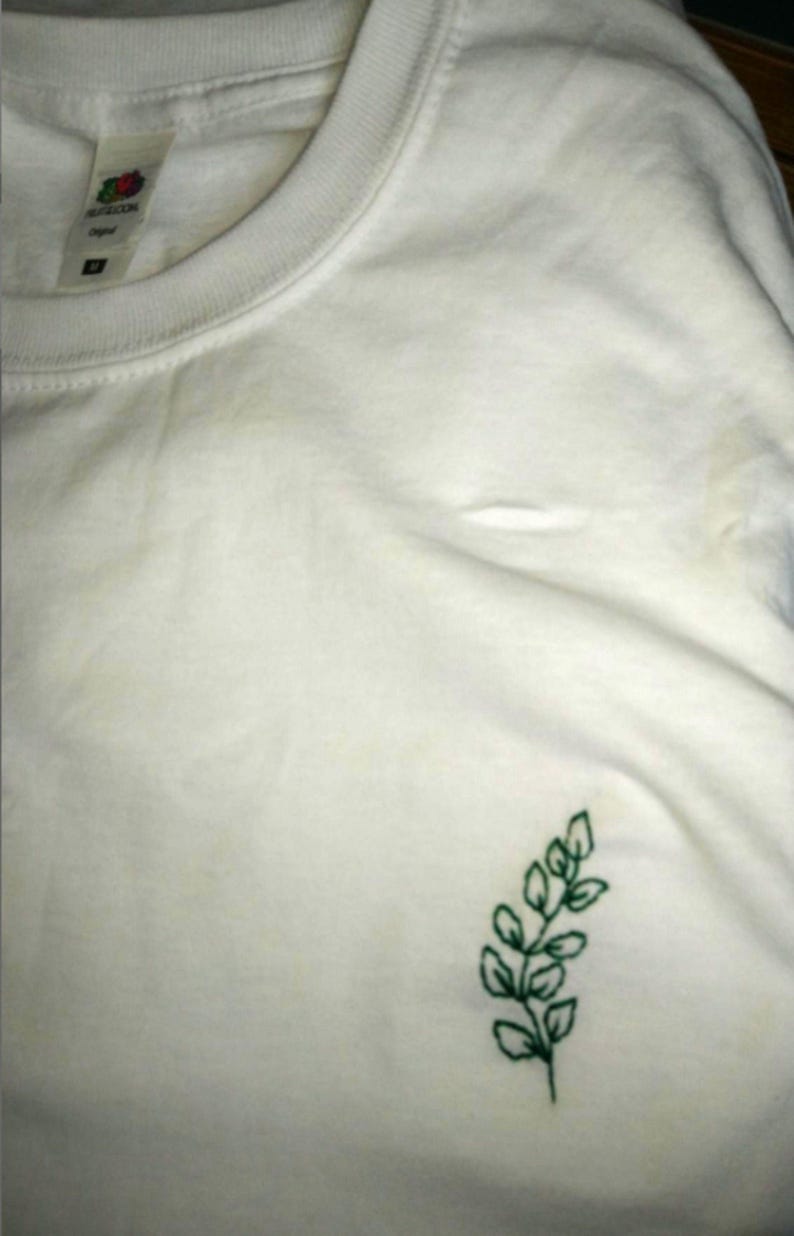 Puede incluir: Camiseta blanca con cuello redondo y mangas cortas. Un peque&ntilde;o dise&ntilde;o de hojas verdes bordadas est&aacute; en el pecho izquierdo. La camiseta est&aacute; hecha de un material suave. La etiqueta de la camiseta es visible.