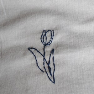 Puede incluir: Tela de color crema con un dise&ntilde;o de tulip&aacute;n bordado en azul oscuro. El tulip&aacute;n se representa con un contorno simple, con una cabeza de flor, un tallo y hojas. El bordado es limpio y detallado.