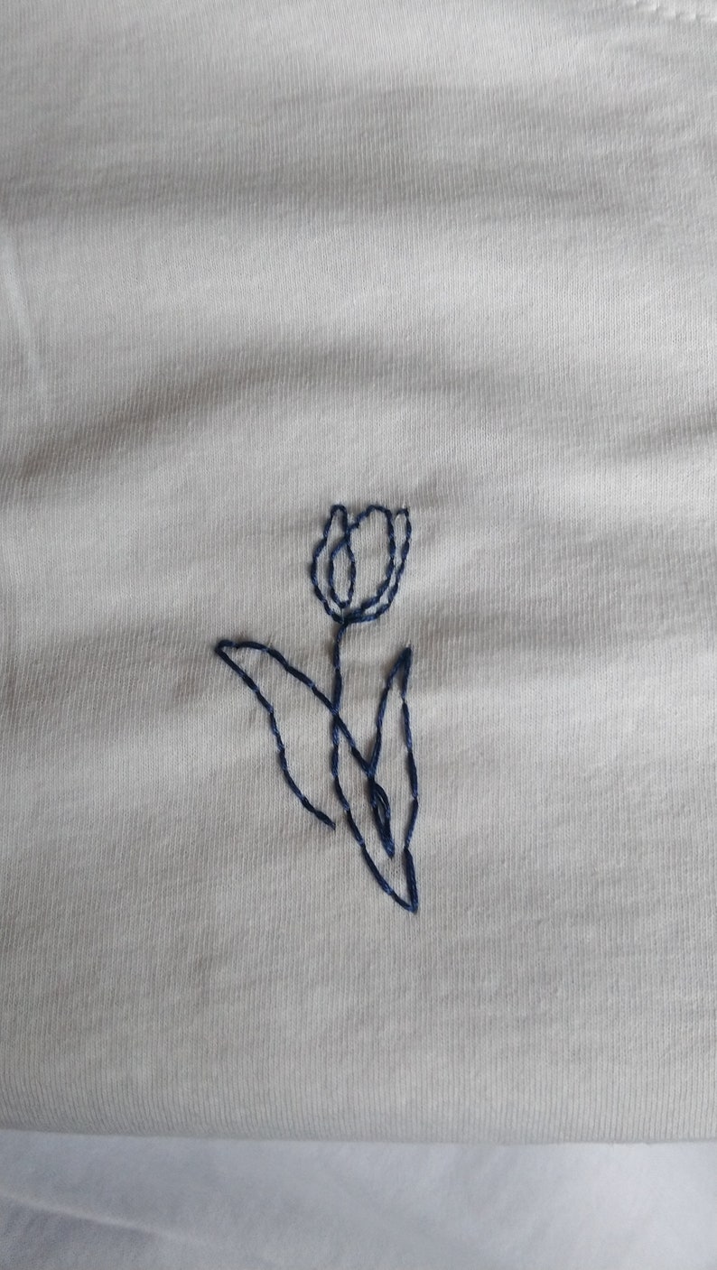 Puede incluir: Tela de color crema con un dise&ntilde;o de tulip&aacute;n bordado en azul oscuro. El tulip&aacute;n se representa con un contorno simple, con una cabeza de flor, un tallo y hojas. El bordado es limpio y detallado.