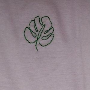 Puede incluir: Una tela de color p&uacute;rpura claro con un dise&ntilde;o de hoja de monstera verde cosida a mano. La hoja est&aacute; centrada y detallada con un contorno simple, agregando un toque de decoraci&oacute;n inspirada en la naturaleza.