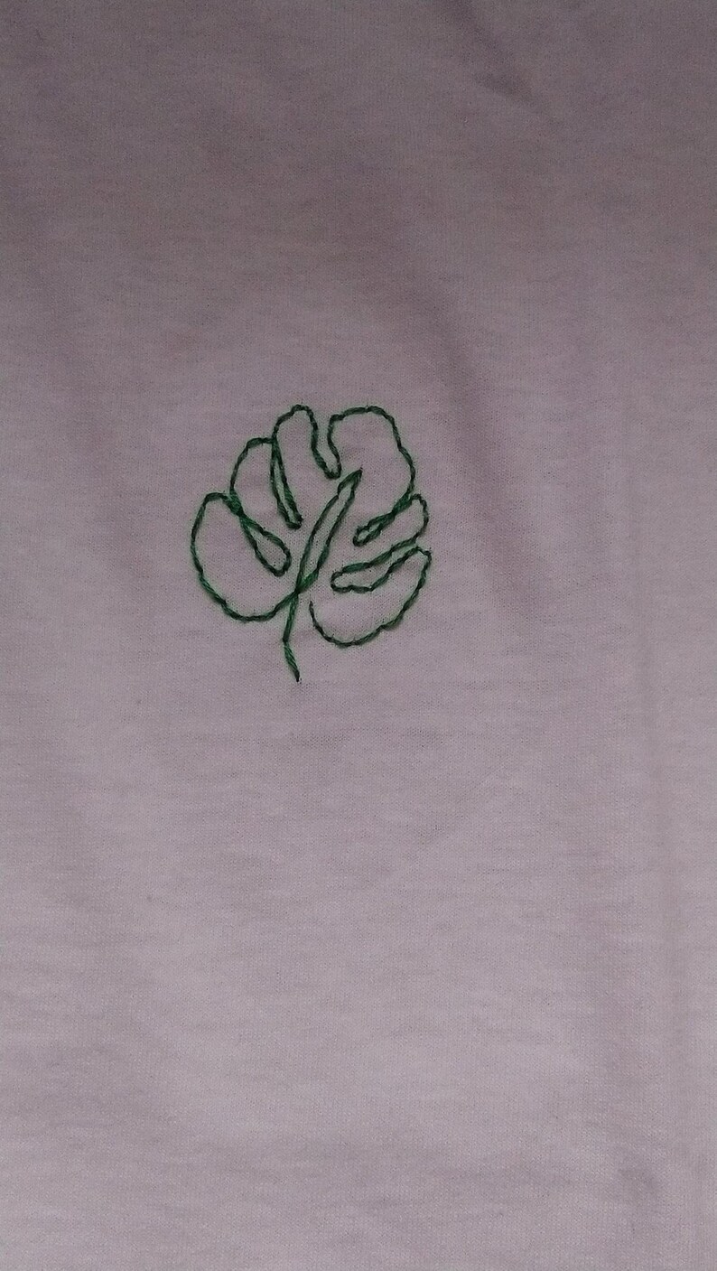 Puede incluir: Una tela de color p&uacute;rpura claro con un dise&ntilde;o de hoja de monstera verde cosida a mano. La hoja est&aacute; centrada y detallada con un contorno simple, agregando un toque de decoraci&oacute;n inspirada en la naturaleza.