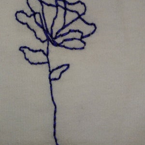 Puede incluir: Dise&ntilde;o de bordado floral cosido a mano. La imagen presenta un contorno azul de una rosa sobre un fondo de tela blanca. La flor tiene m&uacute;ltiples p&eacute;talos y un tallo con dos hojas. El bordado est&aacute; hecho con un solo hilo.
