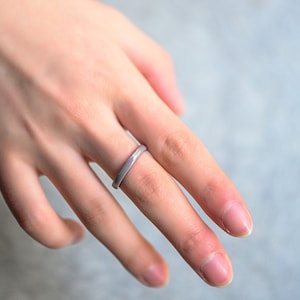 Puede incluir: Un anillo simple de color plateado con un diseño sutil ligeramente elevado. El anillo se lleva en el tercer dedo de la mano izquierda.