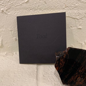 Puede incluir: Una caja cuadrada negra con la palabra "Tool" en relieve en la parte delantera. Un trozo de piedra oscura y pulida con vetas marrones y negras se encuentra a la derecha de la caja. El fondo es una pared de ladrillos blancos.