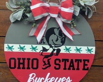Op de afbeelding: Ronde houten bord met het logo en de tekst van de Ohio State Buckeyes, met een rood-witte gestreepte strik en groen. Het bord bevat de woorden "OHIO STATE Buckeyes" in vette letters.