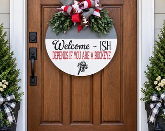 May include: A round wooden door sign featuring a grey and white background with the text "Welcome - ISH DEPENDS IF YOU ARE A BUCKEYE" in black and red, accented by a decorative bow with football patterns and green foliage at the top.