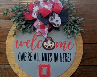 May include: A round wooden door sign with a gray center featuring the red text "welcome" and black text "(WE'RE ALL NUTS IN HERE)" above a red block letter O. A large bow with football and red plaid patterns sits atop with green faux foliage.