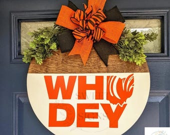 Pode incluir: Um pendurador de porta redondo de madeira com fundo branco e as palavras "WHO DEY" em letras vermelhas. O pendurador é decorado com um laço preto e laranja e folhagem verde.