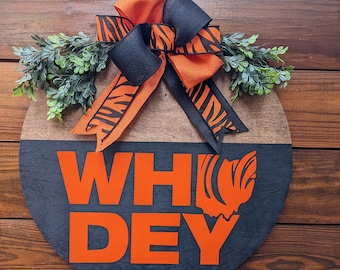 May include: A circular wooden sign with the words "WHO DEY" in orange lettering. The top half is natural wood, the bottom half is dark grey. An orange and black tiger-striped bow and greenery are at the top.
